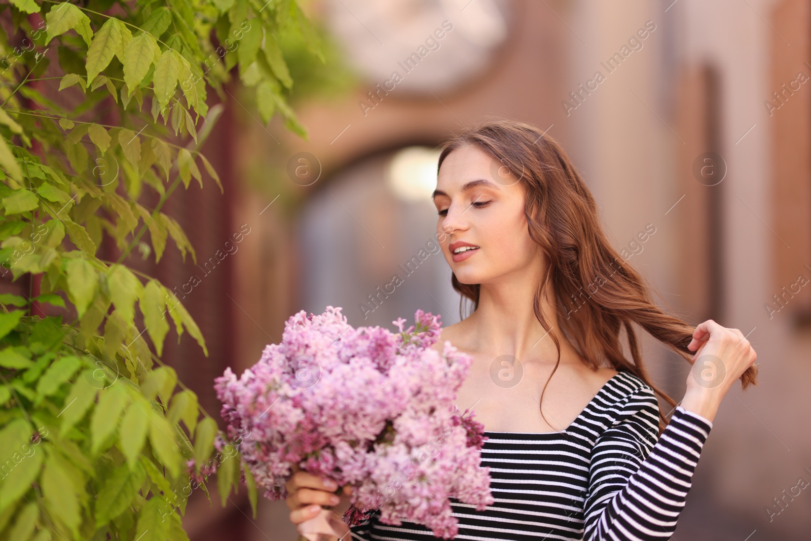 Smiling woman with bouquet of lilac flowers near tree on city street Photo of Smiling woman with bouquet of lilac flowers near tree on city street