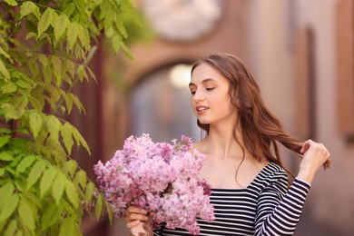 Photo of Smiling woman with bouquet of lilac flowers near tree on city street
