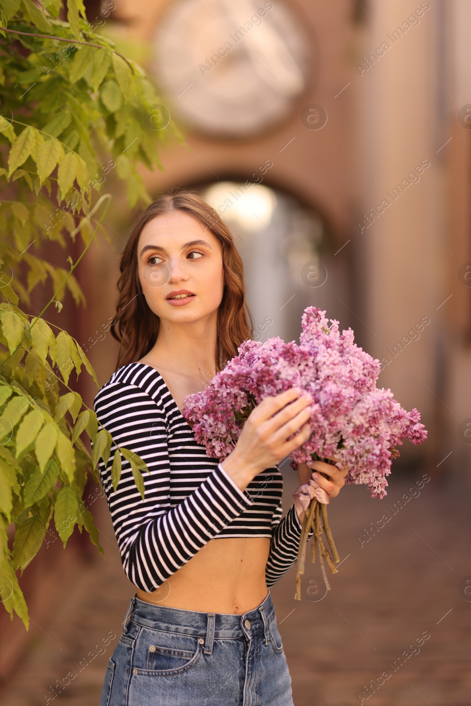 Beautiful woman with bouquet of lilac flowers near tree on city street Photo of Beautiful woman with bouquet of lilac flowers near tree on city street