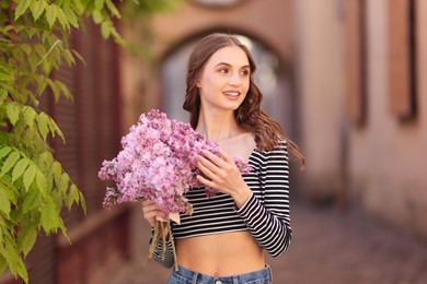 Photo of Smiling woman with bouquet of lilac flowers near tree on city street