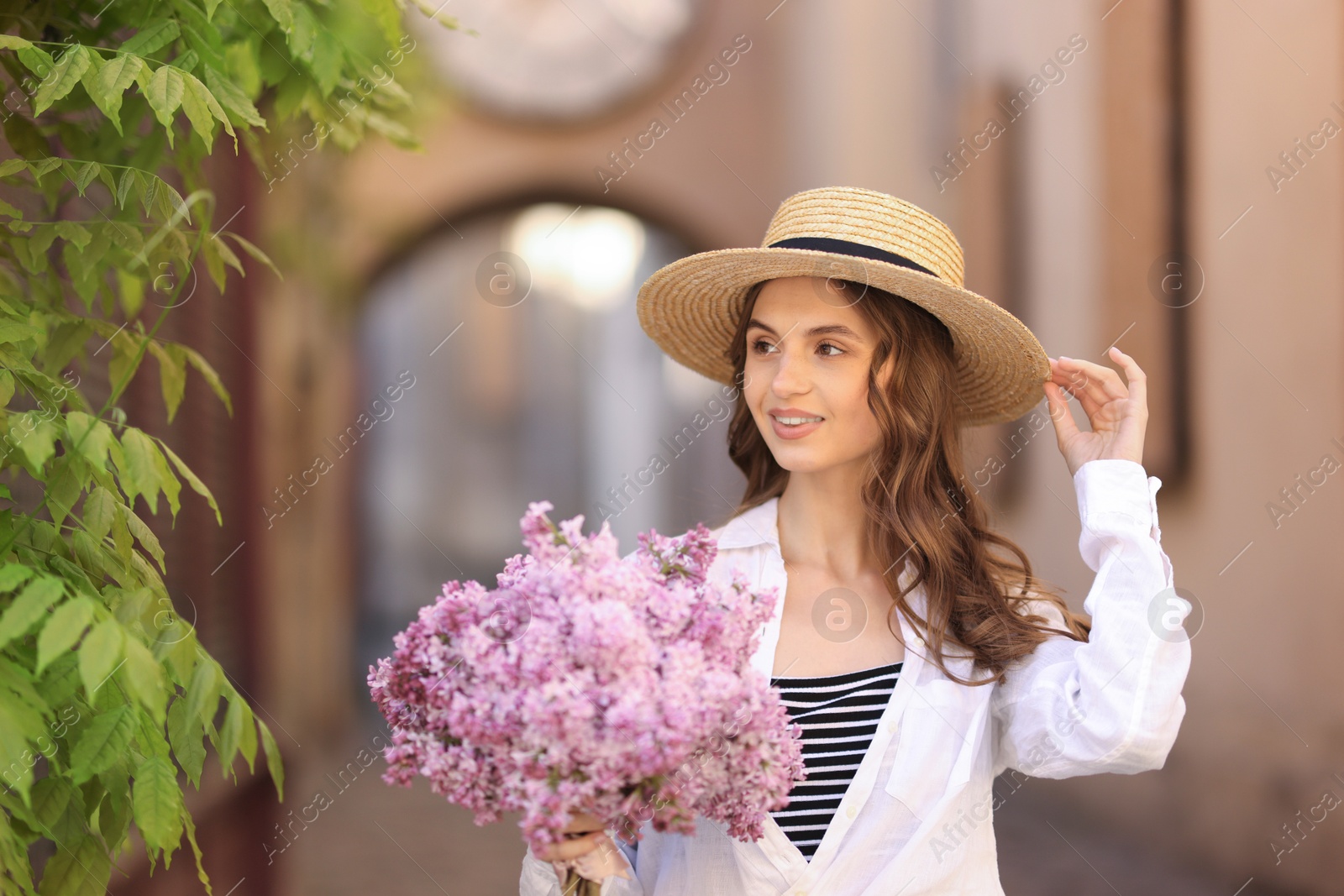 Smiling woman in hat with bouquet of lilac flowers near tree on city street Photo of Smiling woman in hat with bouquet of lilac flowers near tree on city street