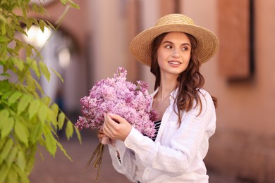 Photo of Smiling woman in hat with bouquet of lilac flowers near tree on city street