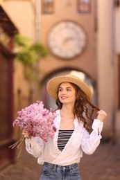 Photo of Smiling woman in hat with bouquet of lilac flowers on city street