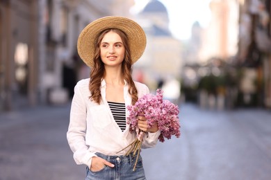 Photo of Smiling woman in hat with bouquet of lilac flowers on city street