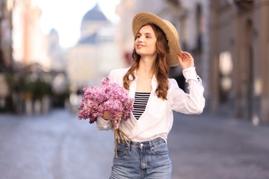 Photo of Smiling woman in hat with bouquet of lilac flowers on city street