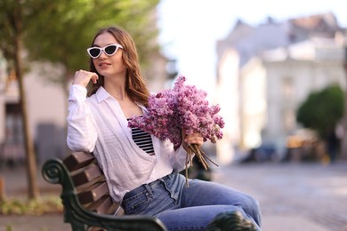 Photo of Smiling woman in sunglasses with bouquet of lilac flowers sitting on bench outdoors