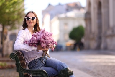 Photo of Smiling woman in sunglasses with bouquet of lilac flowers sitting on bench outdoors. Space for text