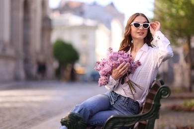Photo of Smiling woman in sunglasses with bouquet of lilac flowers sitting on bench outdoors. Space for text