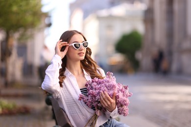 Photo of Beautiful woman in sunglasses with bouquet of lilac flowers sitting on bench outdoors