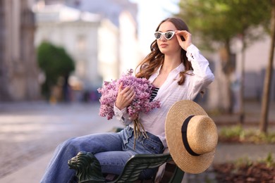 Photo of Beautiful woman in sunglasses with bouquet of lilac flowers sitting on bench outdoors