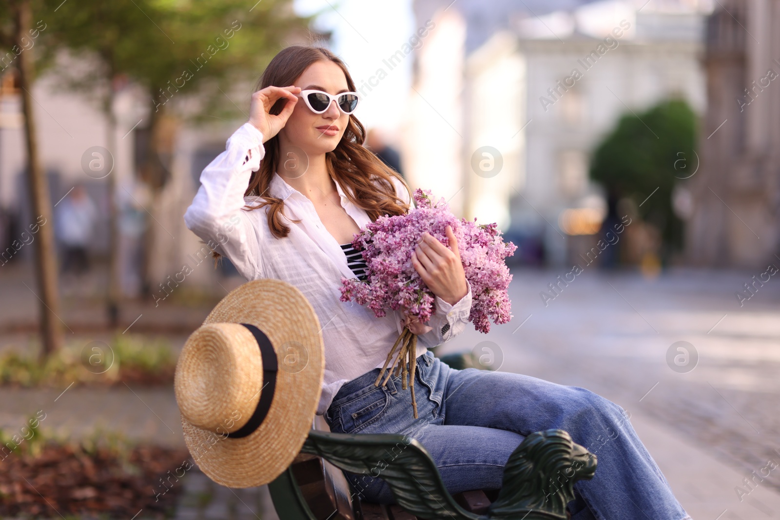 Beautiful woman in sunglasses with bouquet of lilac flowers sitting on bench outdoors Photo of Beautiful woman in sunglasses with bouquet of lilac flowers sitting on bench outdoors