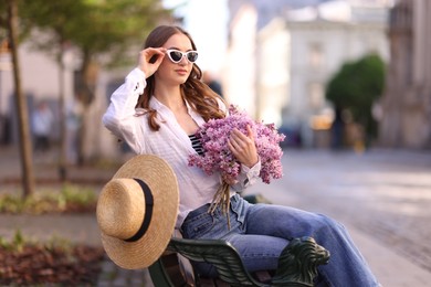 Photo of Beautiful woman in sunglasses with bouquet of lilac flowers sitting on bench outdoors