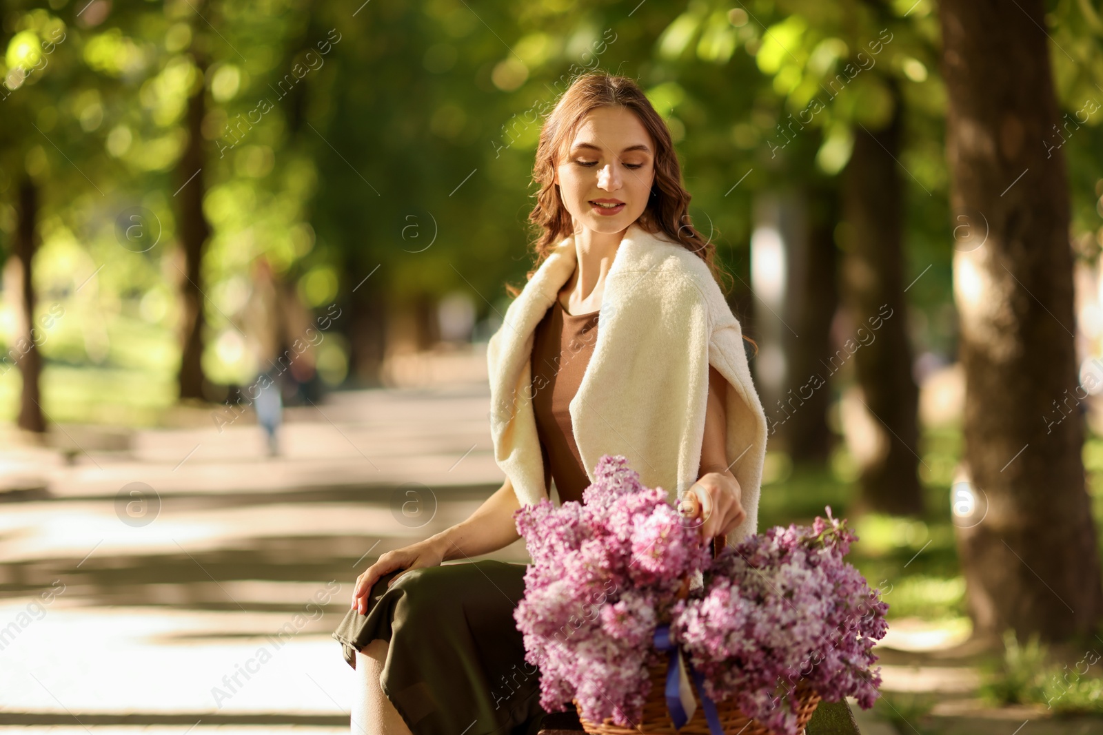 Smiling woman with basket of lilac flowers sitting on bench outdoors in morning. Space for text Photo of Smiling woman with basket of lilac flowers sitting on bench outdoors in morning. Space for text