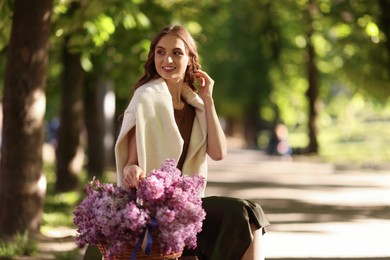Photo of Smiling woman with basket of lilac flowers sitting on bench outdoors in morning. Space for text