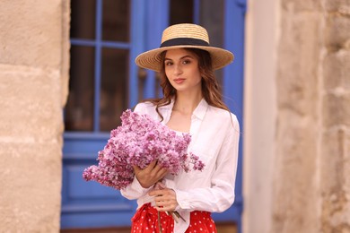 Photo of Beautiful woman in hat with bouquet of lilac flowers near building outdoors