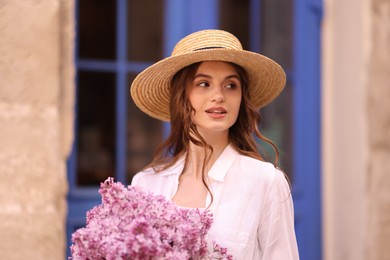 Photo of Beautiful woman in hat with bouquet of lilac flowers near building outdoors