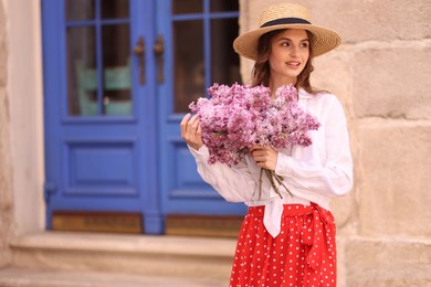 Photo of Smiling woman in hat with bouquet of lilac flowers near building outdoors. Space for text