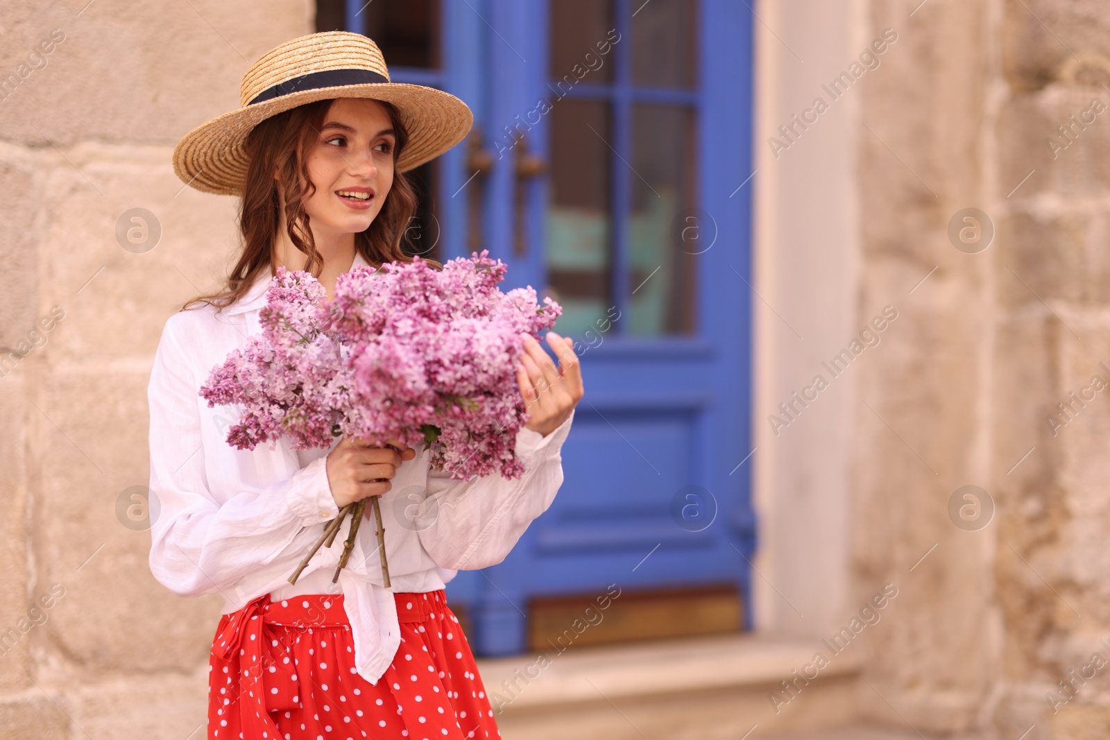 Smiling woman in hat with bouquet of lilac flowers near building outdoors. Space for text Photo of Smiling woman in hat with bouquet of lilac flowers near building outdoors. Space for text