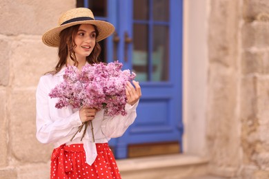 Photo of Smiling woman in hat with bouquet of lilac flowers near building outdoors. Space for text