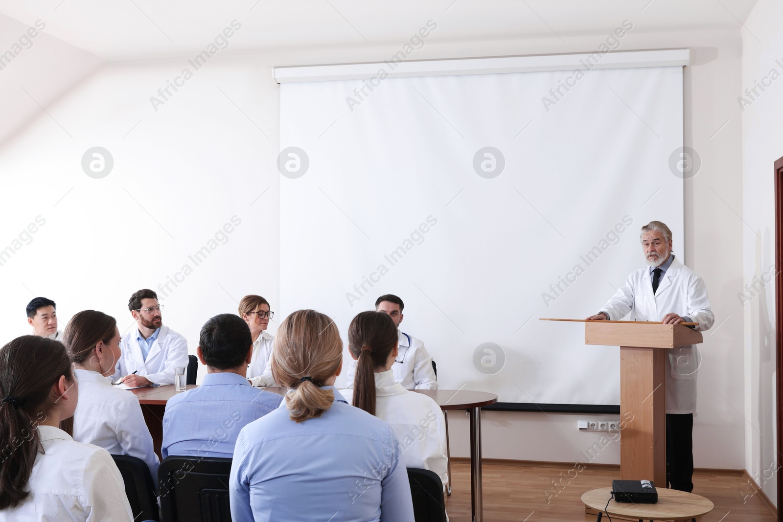 Senior doctor giving lecture in conference room with projection screen Photo of Senior doctor giving lecture in conference room with projection screen
