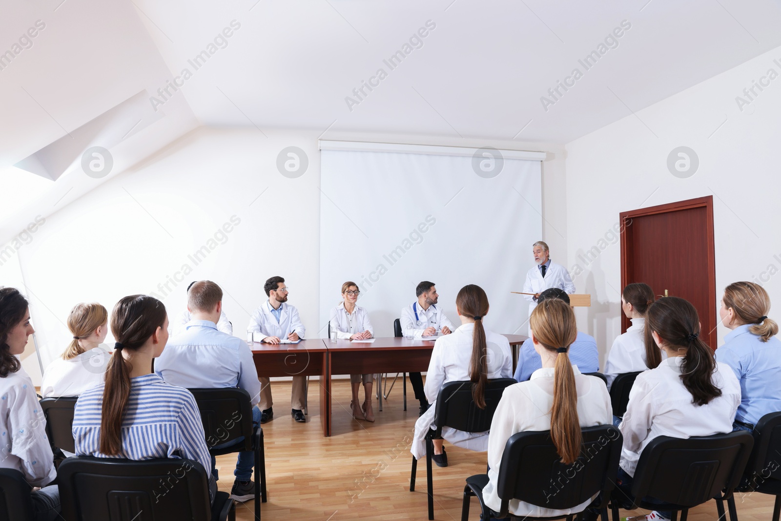 Senior doctor giving lecture in conference room with projection screen Photo of Senior doctor giving lecture in conference room with projection screen