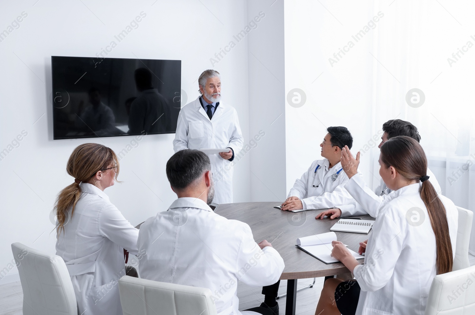 Team of doctors listening to senior speaker report near tv screen in meeting room. Medical conference Photo of Team of doctors listening to senior speaker report near tv screen in meeting room. Medical conference