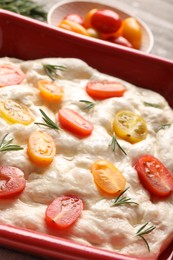 Photo of Raw focaccia with tomatoes and rosemary in baking dish on table, closeup