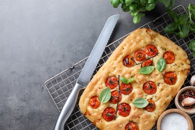 Photo of Delicious focaccia with tomatoes, spices and knife on gray textured table, flat lay. Space for text