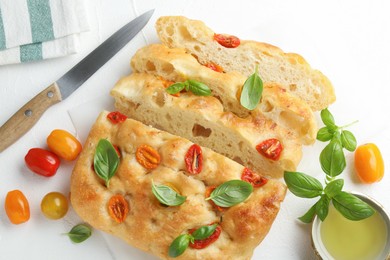 Photo of Cut delicious focaccia with tomatoes, basil, oil and knife on light table, flat lay
