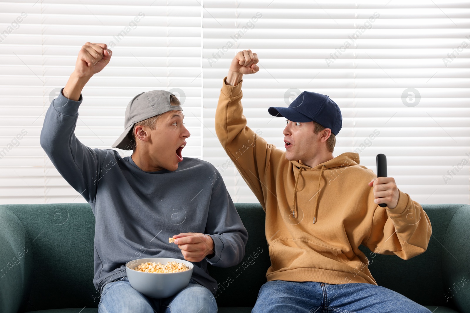 Emotional sports fans with remote control and popcorn watching game together on sofa indoors Photo of Emotional sports fans with remote control and popcorn watching game together on sofa indoors