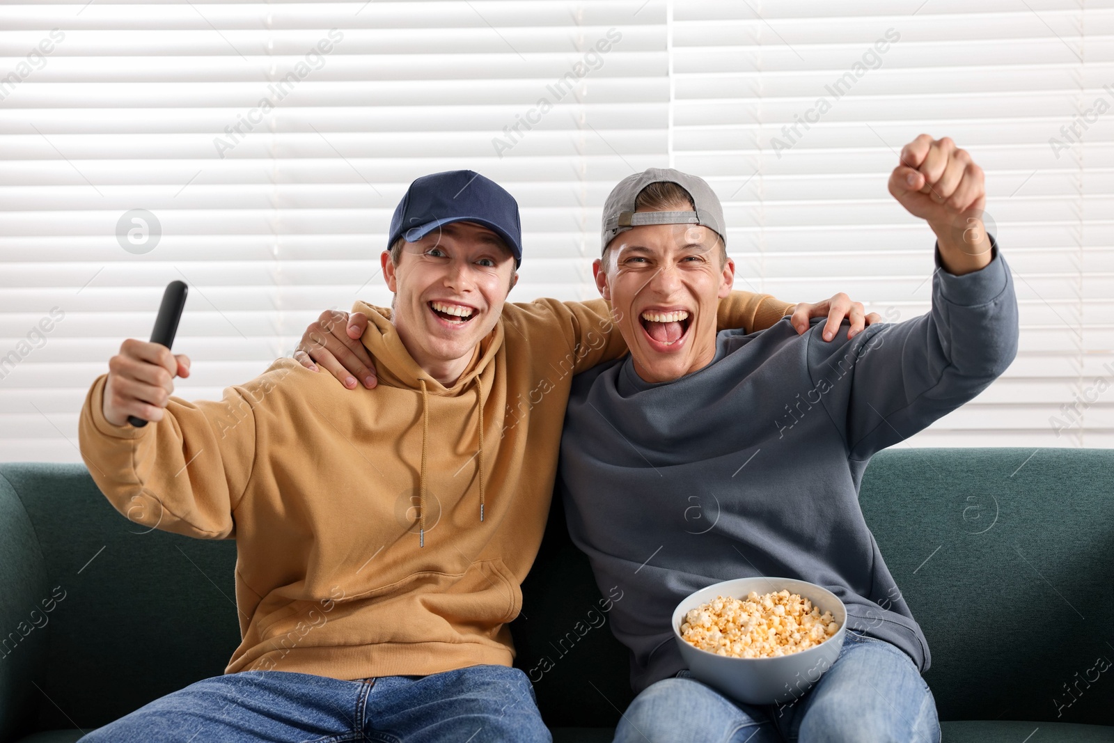 Emotional sports fans with remote control and popcorn watching game together on sofa indoors Photo of Emotional sports fans with remote control and popcorn watching game together on sofa indoors