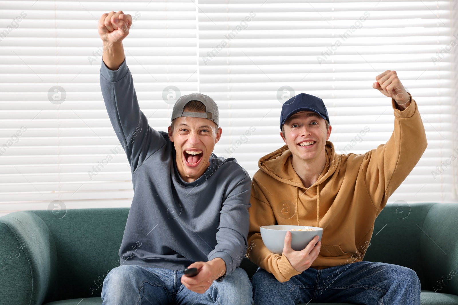 Emotional sports fans with remote control and popcorn watching game together on sofa indoors Photo of Emotional sports fans with remote control and popcorn watching game together on sofa indoors