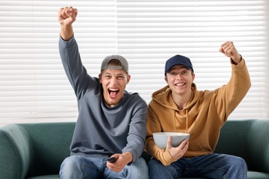 Photo of Emotional sports fans with remote control and popcorn watching game together on sofa indoors