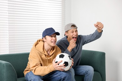 Photo of Emotional sports fans with soccer ball watching game together on sofa indoors