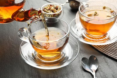 Photo of Pouring herbal tea from teapot into cup at black table, closeup