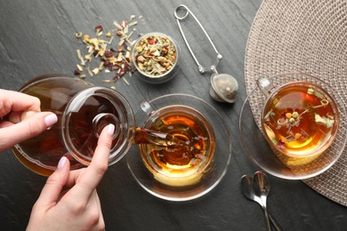 Photo of Woman pouring herbal tea from teapot into cup at black table, top view