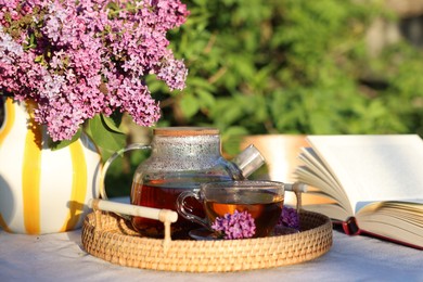 Photo of Beautiful lilac flowers, tea and book on table in garden, closeup
