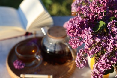 Photo of Beautiful lilac flowers, tea and book on table in garden, closeup