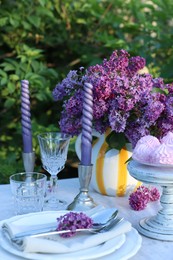 Photo of Beautiful lilac flowers, zephyrs, plates, cutlery, glasses and candles on table in garden, closeup