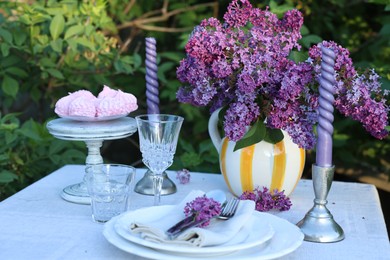 Photo of Beautiful lilac flowers, zephyrs, plates, cutlery, glasses and candles on table in garden