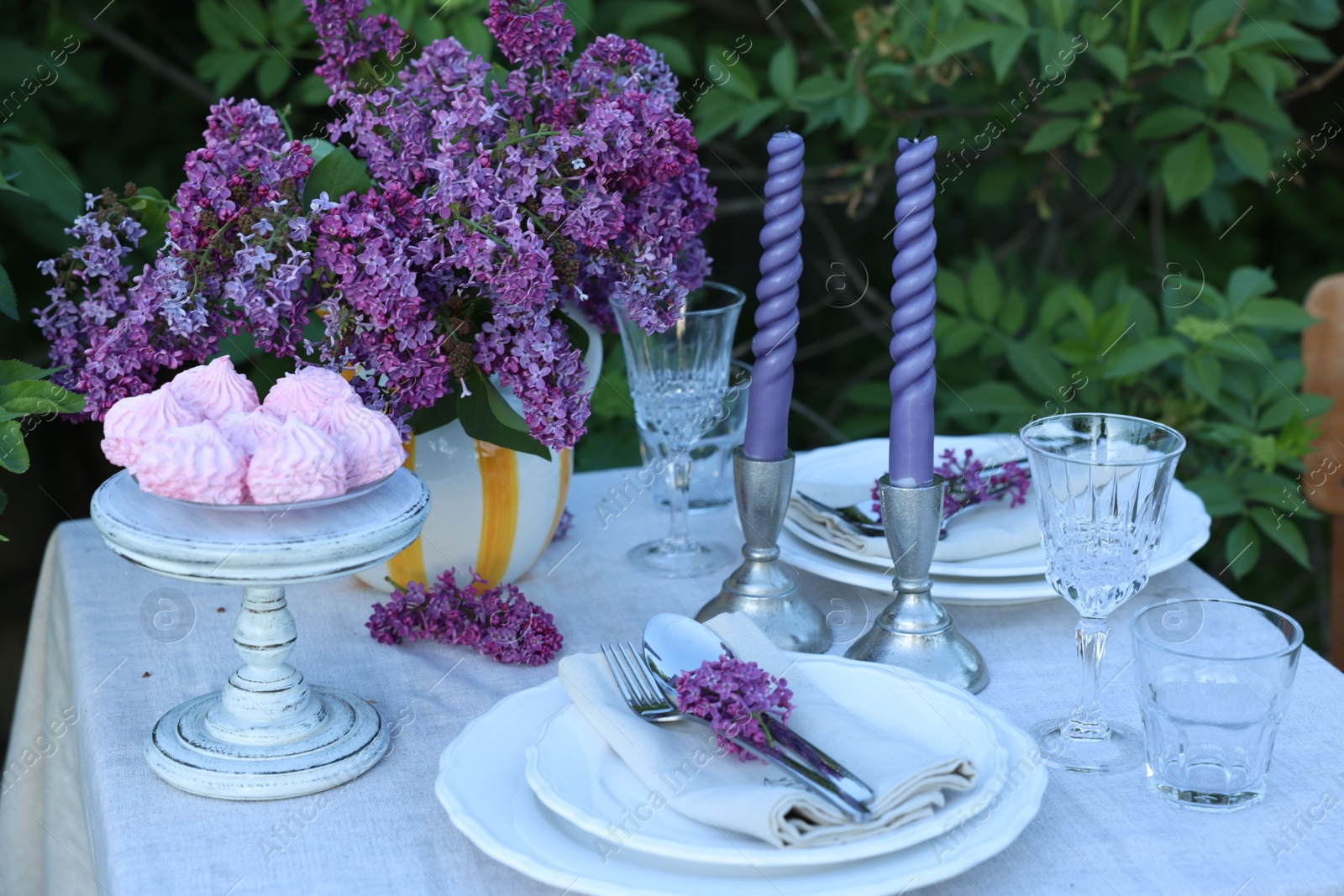Photo of Beautiful lilac flowers, zephyrs, plates, cutlery, glasses and candles on table in garden