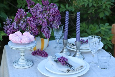 Photo of Beautiful lilac flowers, zephyrs, plates, cutlery, glasses and candles on table in garden