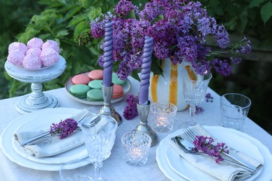 Photo of Beautiful lilac flowers, sweets, plates, cutlery, glasses and candles on table in garden