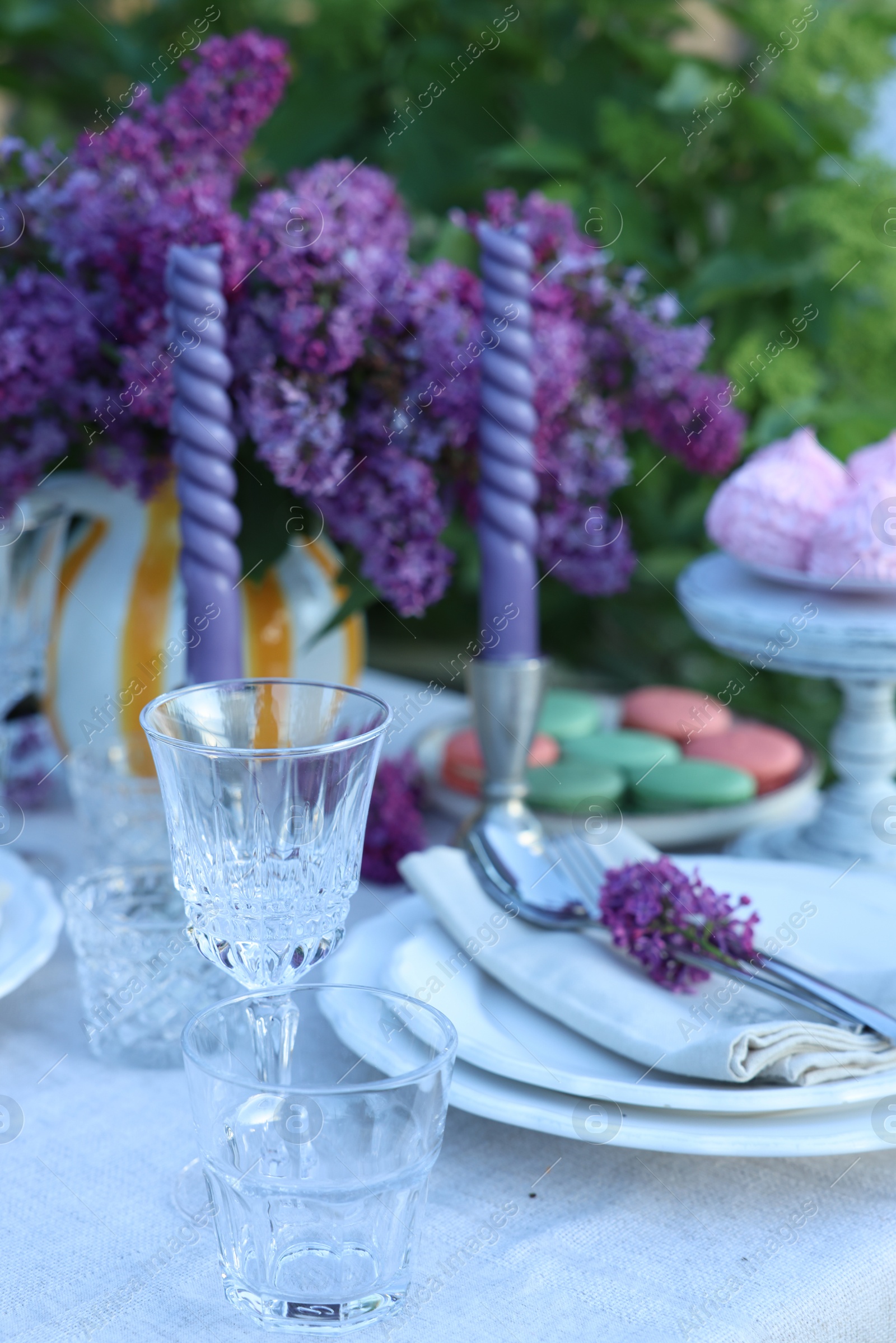 Beautiful lilac flowers, sweets, plates, cutlery, glasses and candles on table in garden, closeup Photo of Beautiful lilac flowers, sweets, plates, cutlery, glasses and candles on table in garden, closeup
