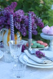 Photo of Beautiful lilac flowers, sweets, plates, cutlery, glasses and candles on table in garden, closeup