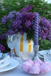 Photo of Beautiful lilac flowers, zephyrs, plates, cutlery, glass and candles on table in garden, closeup