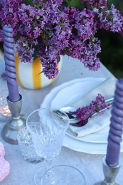 Photo of Beautiful lilac flowers, plates, cutlery, glass and candles on table in garden, above view
