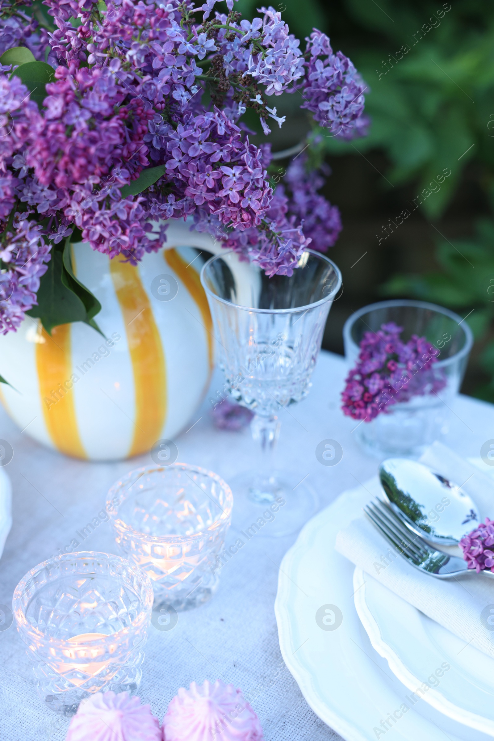 Beautiful lilac flowers, plates, cutlery, glasses and burning candles on table in garden, closeup Photo of Beautiful lilac flowers, plates, cutlery, glasses and burning candles on table in garden, closeup