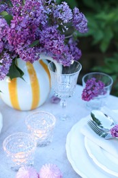 Photo of Beautiful lilac flowers, plates, cutlery, glasses and burning candles on table in garden, closeup