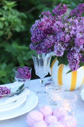 Photo of Beautiful lilac flowers, zephyrs, plates, cutlery, glasses and burning candles on table in garden, closeup